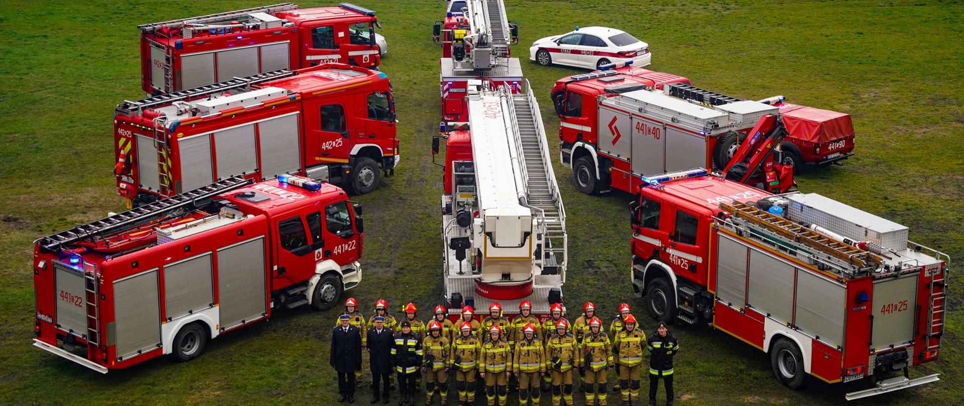 Czerwono srebrne pojazdy strażackie ustawione w kształcie choinki na trawiastym terenie przy rzece. Przed nimi stoją w dwuszeregu strażacy.
