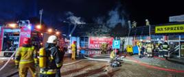 Na wprost znajduje się spalony budynek handlowy "Panelowo". Do jego lewego boku przylega restauracja "Tonic Bar". Po lewej stronie zdjęcia stoi wóz strażacki na sygnałach. Z spalonego dachu unosi się dym. Strażacy biorący udział w akcji znajdują się na całym parkingu, przed budynkiem dotkniętym pożarem oraz na dachu restauracji. Na środku parkingu strażak obsługuje pompę.