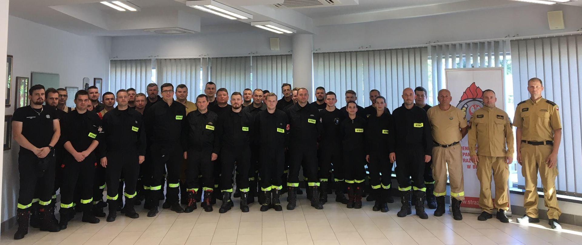 Kilkudziesięciu mężczyzn i kobiet ubranych w czarne ubrania koszarowe strażackie z odblaskowymi paskami, czarnym butami, stoją w Sali konferencyjnej. W tle okna oraz rolety koloru szarego. Ściany koloru białego