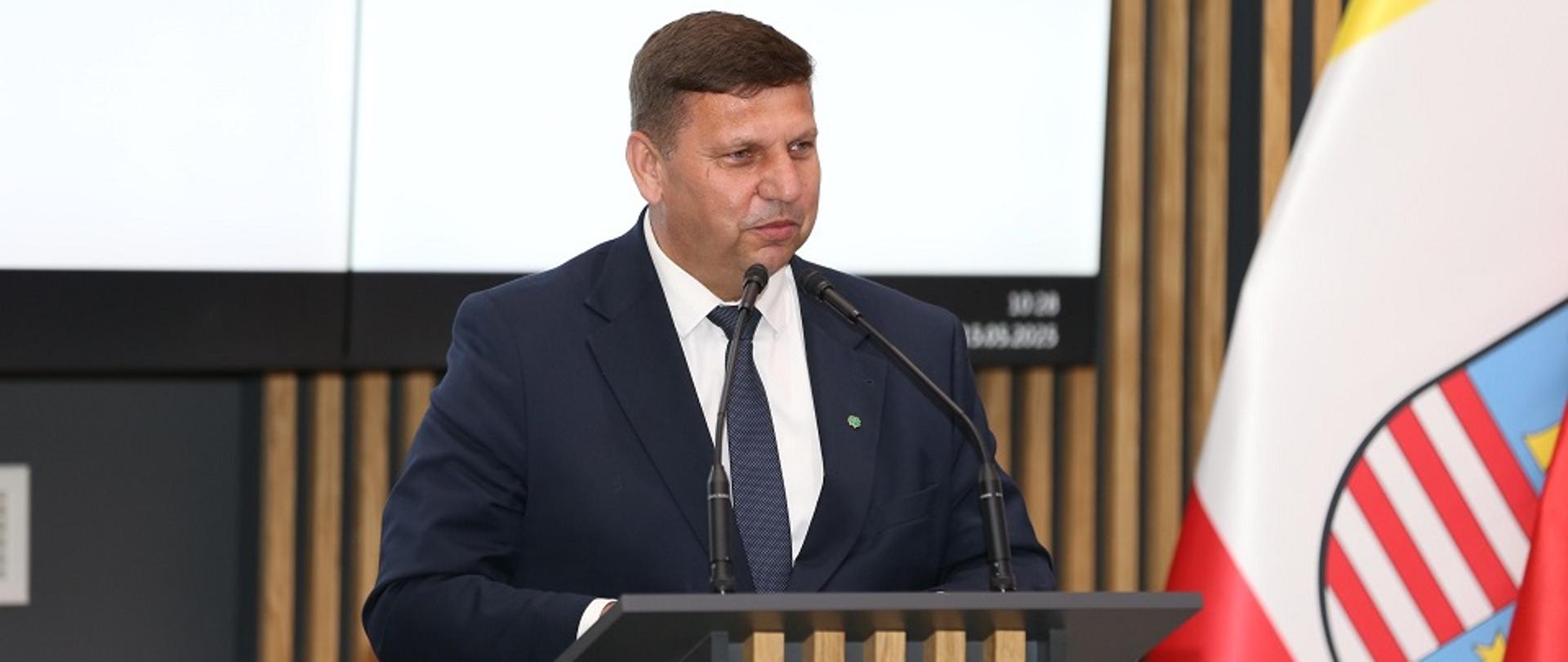 Wicewojewoda Świętokrzyski w granatowym garniturze stoi za mównicą i przy użyciu mikrofonów przemawia do zgromadzonych gości