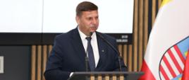 Wicewojewoda Świętokrzyski w granatowym garniturze stoi za mównicą i przy użyciu mikrofonów przemawia do zgromadzonych gości