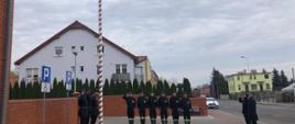 strażacy stoją przed masztem i zostaje podniesiona flaga narodowa na maszt w tle budynki mieszkalne