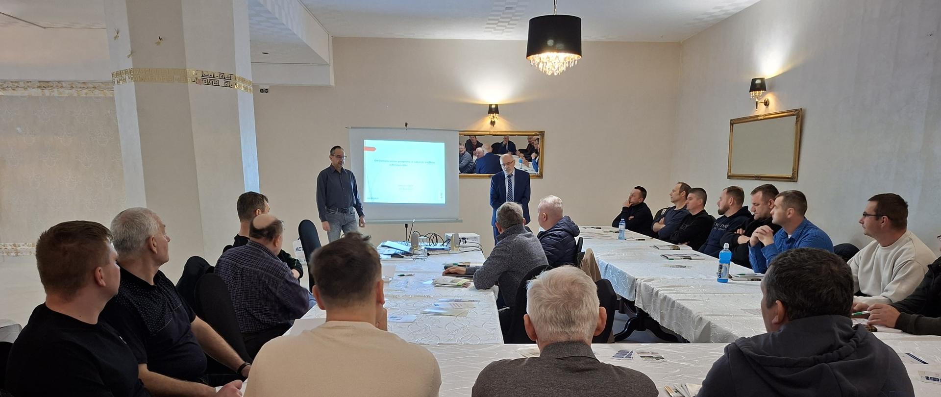 Grupa kilkunastu osób siedzi przy stołach w sali konferencyjnej i słucha dwóch prowadzących stojących przy ekranie z prezentacją wyświetlaną z projektora.