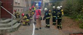 Zdjęcie przedstawia strażaków podczas ćwiczeń ewakuacyjnych w szpitalu.
W tle budynek szpitala i samochody strażackie.
