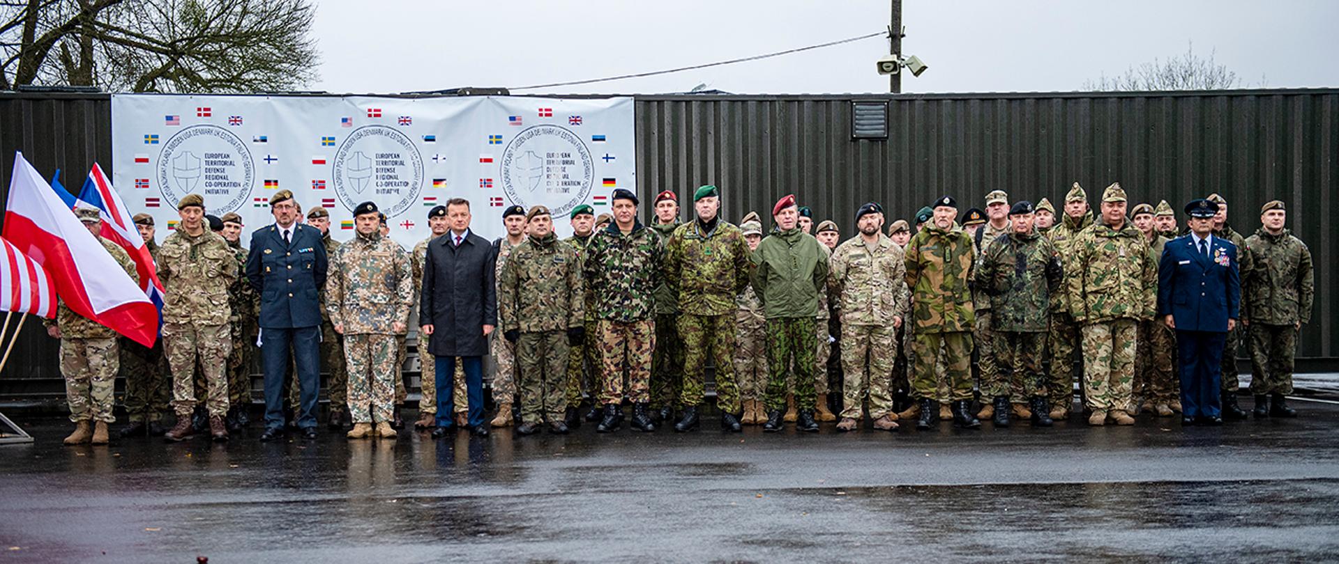 Szef MON z dowódcami WOT innych państwa na placu apelowym w Zegrzu. Po lewej stronie flagi państw. Na górze logo inicjatywy WOT.