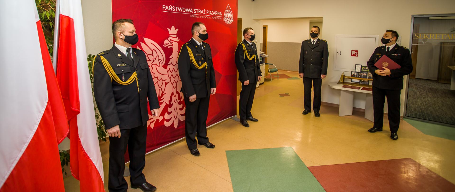 Na zdjęciu widocznych pięciu strażaków w ubraniach galowych z czarnymi maseczkami na twarzach. Po lewej stronie widoczne trzy flagi Polski. Następnie stoi trzech strażaków (kpt. Roman Felusiak, kpt. Roman Wyszka i st. kpt. Krzysztof Letki) w odległości około 1,5 metra od siebie i twarzami skierowani na dwóch pozostałych strażaków, stojących pod kątem około 90 stopni od nich. Od lewej stoi st. bryg. Arkadiusz Kuśmierski a następnie st. bryg. Krzysztof Kędryk (trzymający w ręce ozdobną teczkę koloru czerwonego).