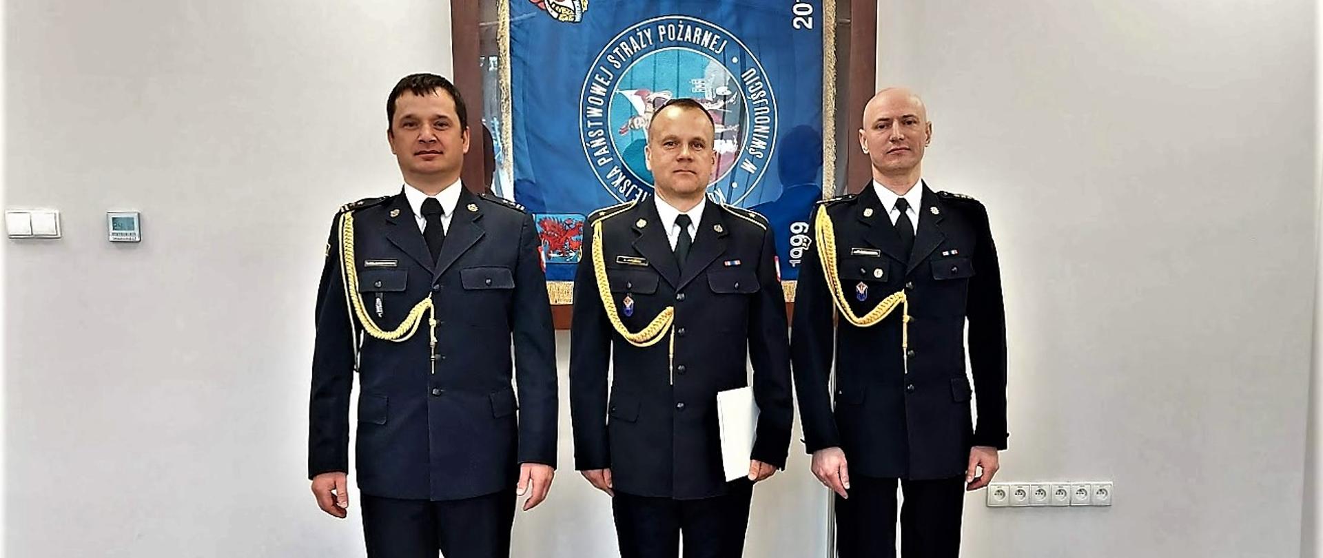 Trzech funkcjonariuszy w mundurach wyjściowych ze sznurem na tle sztandaru komendy w gablocie. Od lewej Zastępca Komendanta Miejskiego, po środku asp. Artur Jakubiak, po prawej Komendant Miejski PSP. 
