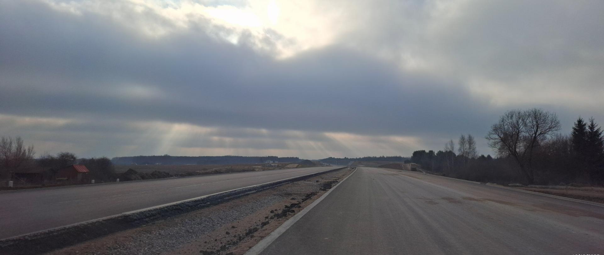 Dwie budowane jezdnie drogi ekspresowej S19, z nawierzchnią asfaltową, puste, na horyzoncie promienie słońce przebijające się przez chmury.