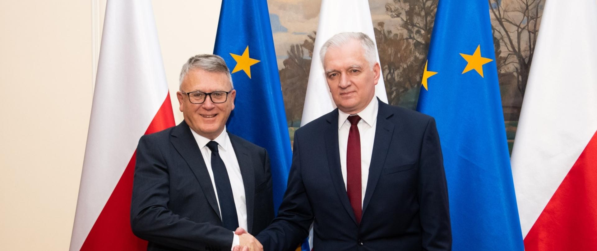 Minister Gowin i komisarz Nicolas Schmit podają sobie ręce, w tle flagi Polski i UE