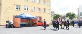 Zdjęcie przedstawia wejście po mszy świętej na plac pododdziału podczas uroczystości Powiatowych Obchodów Dnia Strażaka w KP PSP w Oleśnie. Na pierwszym planie widoczny maszerujący dowódca pododdziału, poczet sztandarowy KP PSP w Oleśnie i klasy mundurowej, strażacy w galowych mundurach i zaproszeni goście. Po lewej stronie stoi namiot promocyjny obrony cywilnej oraz pojazdy ratownicze, w tym quad i czerwony samochód straży pożarnej. W tle znajduje się budynek Komendy oraz inne zabudowania i zieleń.