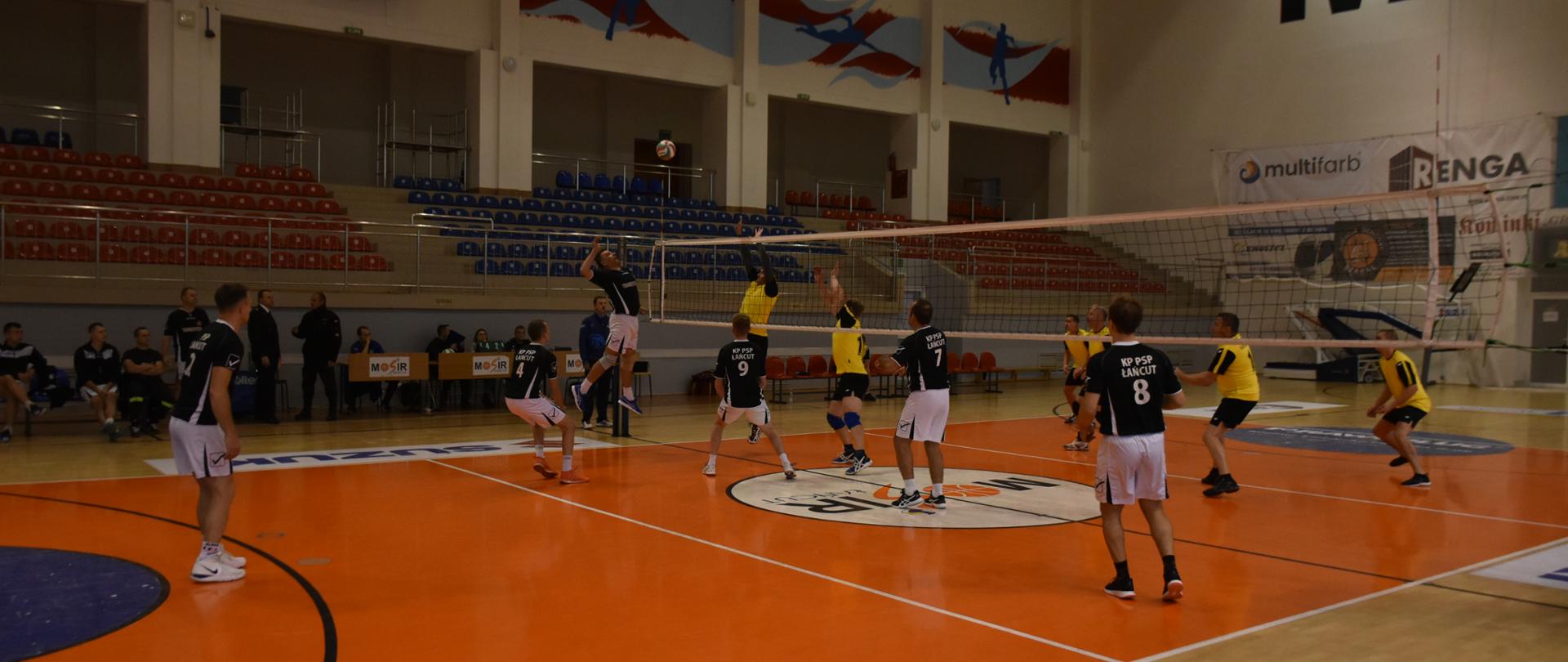 Zdjęcie przedstawia boisko piłki siatkowej w hali MOSiR w Łańcucie. Rozgrywany jest mecz piłki siatkowej pomiędzy reprezentacjami Komendy Powiatowej PSP w Łańcucie i Komendy Powiatowej PSP w Ustrzykach Dolnych. Atakujący z Łańcuta uderza piłkę na lewym skrzydle, dwóch siatkarzy z Ustrzyk Dolnych blokuje atak. W tle stoliki sędziowskie i ławki zawodników rezerwowych.