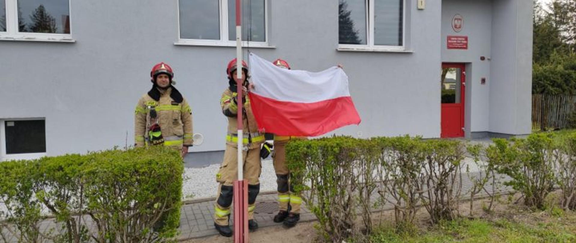 Centralny punkt zdjęcia stanowi masz z flagą i strażacy dokonujący wciągnięcia flagi na maszt. Flaga powiewa od podmuchów wiatru.