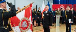 Zdjęcie przedstawia formalną ceremonię odbywającą się w sali gimnastycznej Komendy Wojewódzkiej PSP w Katowicach. Na zdjęciu widoczni są liczni funkcjonariusze w mundurach wyjściowych niektórzy stoją na baczność. Na pierwszym planie poczet sztandarowy odwrócony tyłem: jeden ze strażaków trzyma czerwoną i białą flagę z emblematem i napisem "OJCZYZNA", naprzeciwko stoi śląski komendant wojewódzki st. bryg. Damian Legierski. W tle znajduje się trybuna z zaproszonymi gośćmi więcej osób. Sceneria obejmuje czerwone dywany i wypolerowane podłogi. Za grupą ludzi widoczne są również flagi narodowe.