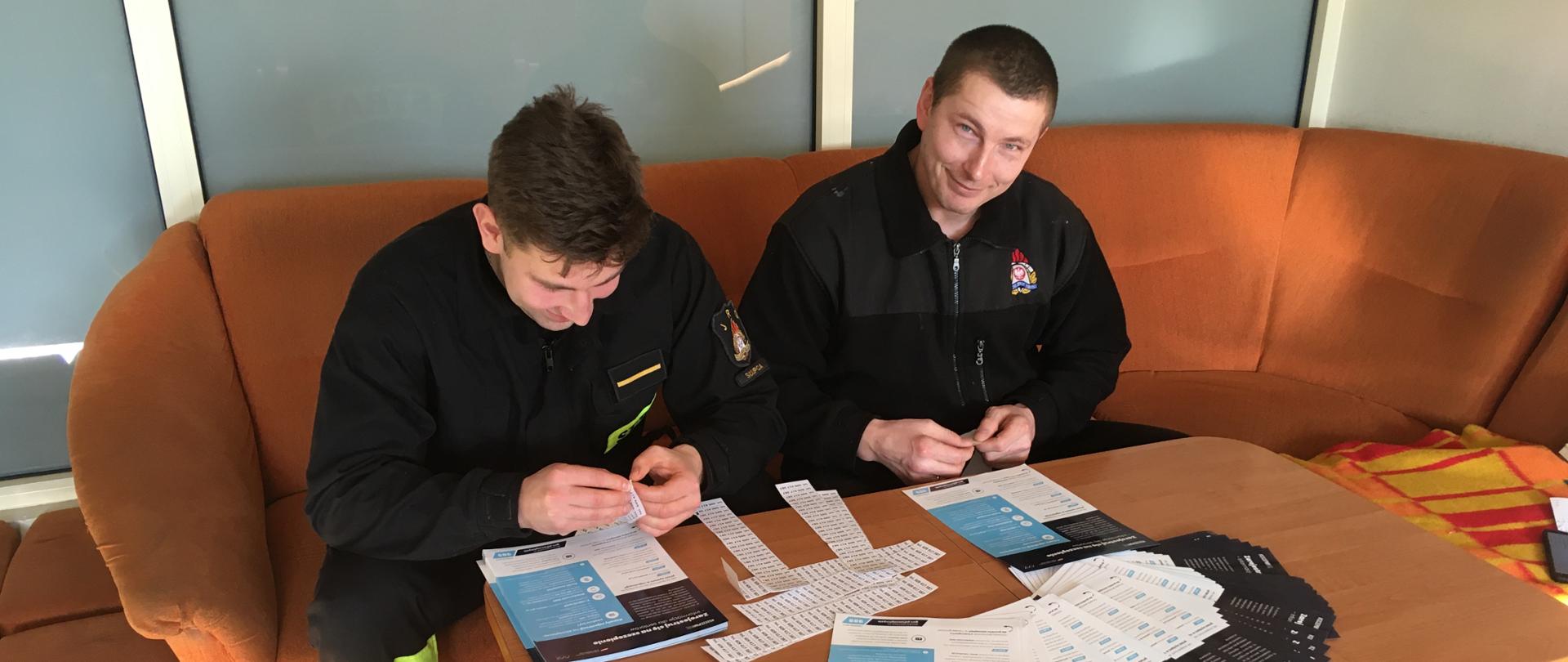 dwóch strażaków siedzących przy stoliku i naklejają numer telefonu na ulotki