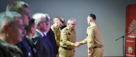 Na zdjęciu widać grupę mężczyzn ustawionych w rzędzie podczas oficjalnej ceremonii. Kilku z nich nosi mundury w kolorze piaskowym z polskimi flagami na rękawach, a inni są ubrani w garnitury. Dwóch mężczyzn na pierwszym planie podaje sobie ręce w geście przywitania. W tle widoczny jest czerwony baner oraz mównica z logo Państwowej Straży Pożarnej.