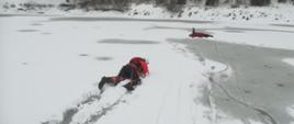zdjęcie przedstawia zamarznięty zbiornik wodny i strażaka, który czołgając się po lodzie idzie na pomoc drugiemu strażakowi