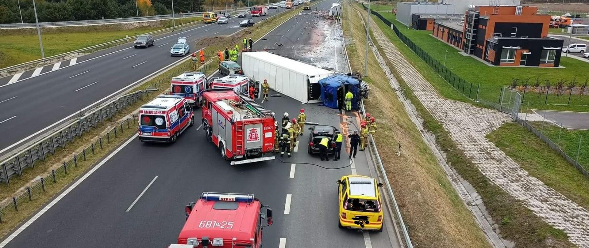 Zdjęcie przedstawia trasę szybkiego ruchu na której doszło do wypadku samochodowego. Na drodze znajdują się dwa czerwone samochody straży pożarnej, trzy karetki pogotowia ratunkowego oraz czarny i żółty samochód osobowy. Obok pojazdów na prawym boku leży samochód ciężarowy, w który uderzył samochód koloru szarego. Widoczni są strażacy, policjanci oraz ratownicy medyczni. W oddali znajduje się uszkodzony samochód ciężarowy oraz osobowy, jezdnia jest pobrudzona na biało. Na przeciwległym pasie ruchu ciągnie się długi korek z samochodów.