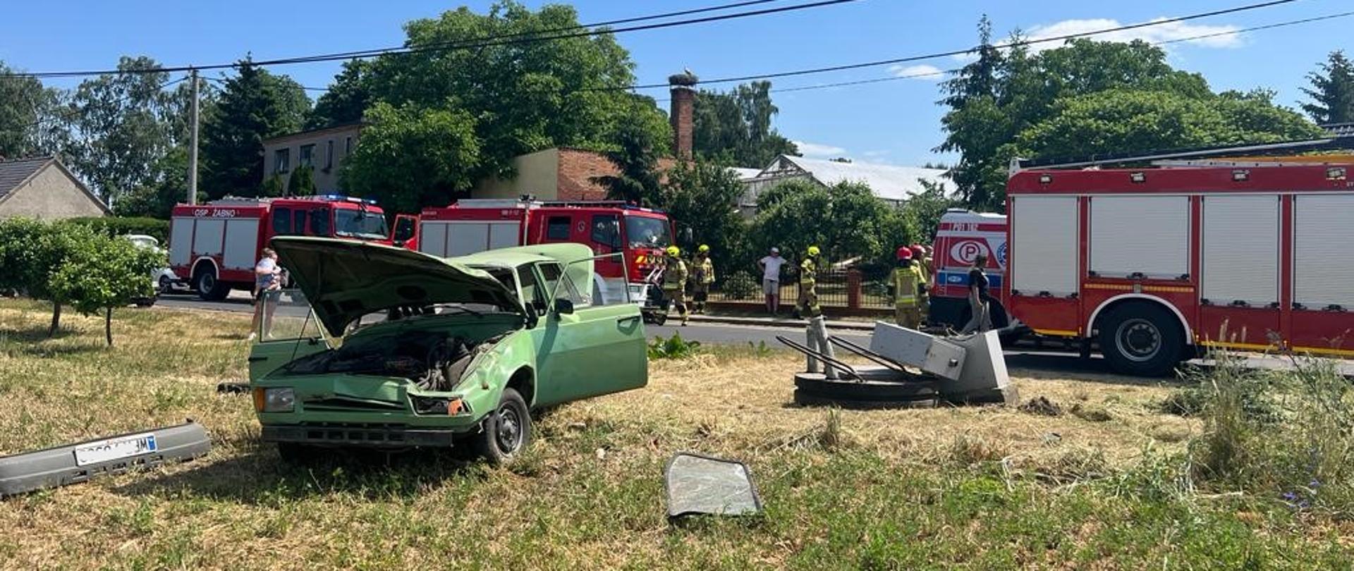 Na zdjęciu czerwone samochody strażackie oraz karetka pogotowia ratunkowego, w tyle strażacy wraz z ratownikami medycznymi udzielający pomocy poszkodowanym, przed nimi wrak pojazdu oraz uszkodzona instalacja przepompowni