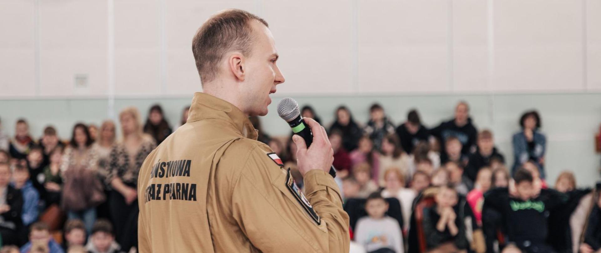 Strażaka, który przemawia do zgromadzonych dzieci. Strażak stoi naprzeciwko tych dzieci. W ręce trzyma mikrofon. Ubrany jest w mundur służbowy. Na plecach widnieje napis: Państwowa Straż Pożarna.