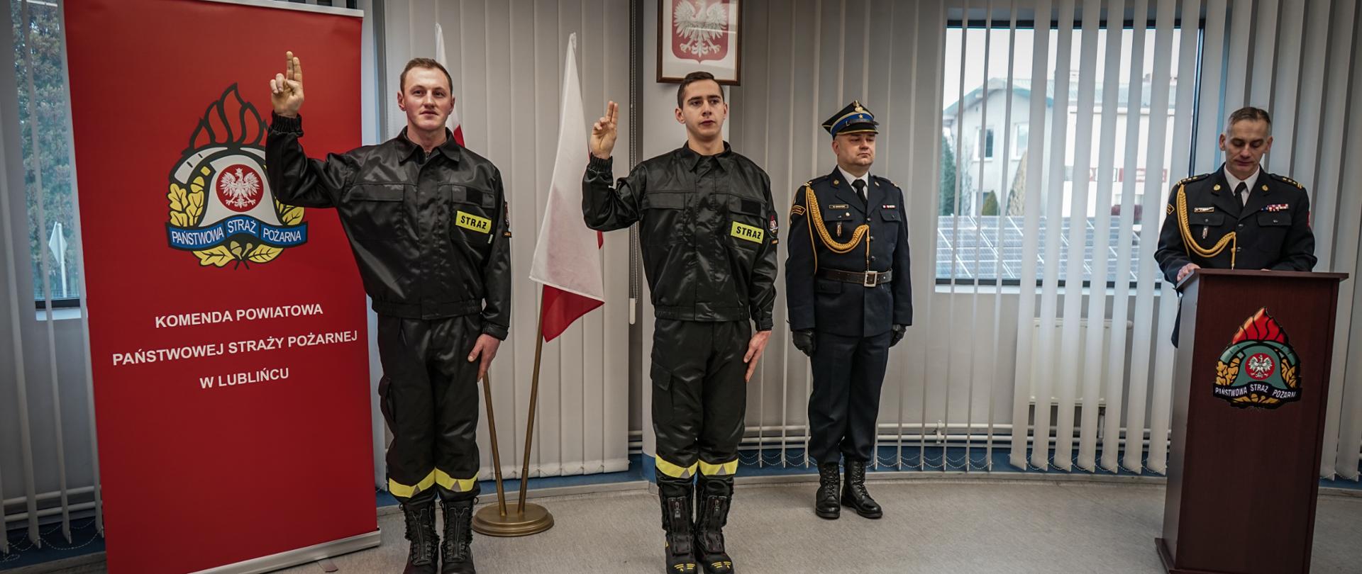 Zdjęcie zrobione w pomieszczeniu strażnicy na tle godła Rzeczpospolitej Polskiej, dwóch Flag Rzeczpospolitej Polskiej oraz po lewej stronie banneru Komendy Powiatowej PSP w Lublińcu. Na pierwszym planie nowoprzyjęci strażacy stojący na baczność i ubrani w umundurowanie koszarowe składają ślubowanie podnosząc prawe ręce, po prawej stronie Komendant w ubraniu galowym stojący za mównicą oraz dowódca uroczystości w umundurowaniu galowym z pasem i w czapce.
