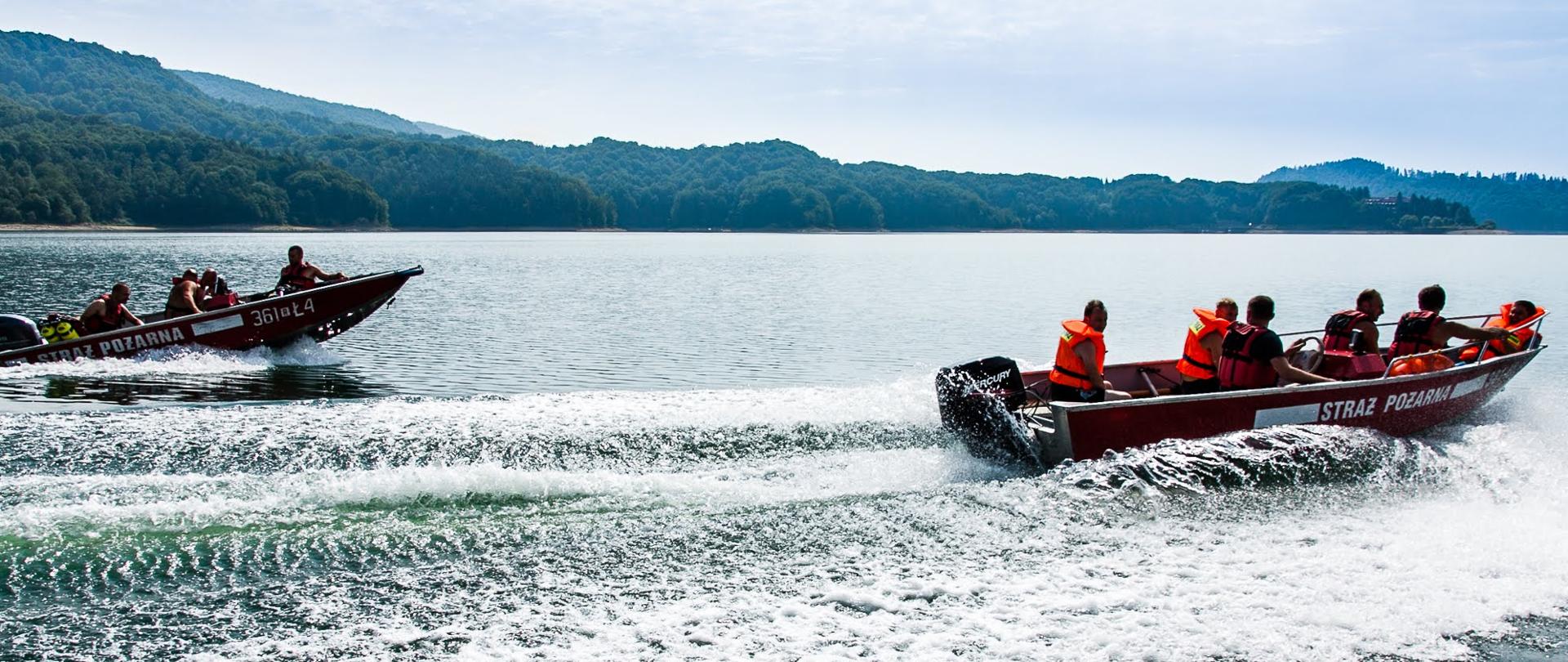 Zdjęcie przedstawia ratowników ze Specjalistycznej Grupy Ratownictwa Wodno - Nurkowego poruszających się dwoma łodziami po jeziorze. W prawej części zdjęcia na łodzi znajduje się 6 strażaków-ratowników w kapokach, jeden z nich kieruje płynącą łódką. Druga z łodzi znajdująca się w tyle do kierunku poruszania się obydwu łodzi transportuje 4 ludzi, z których jeden z nich odpowiada za jej prowadzenie. Na łódkach widoczny sprzęt do nurkowania, na boku napisy Straż pożarna.