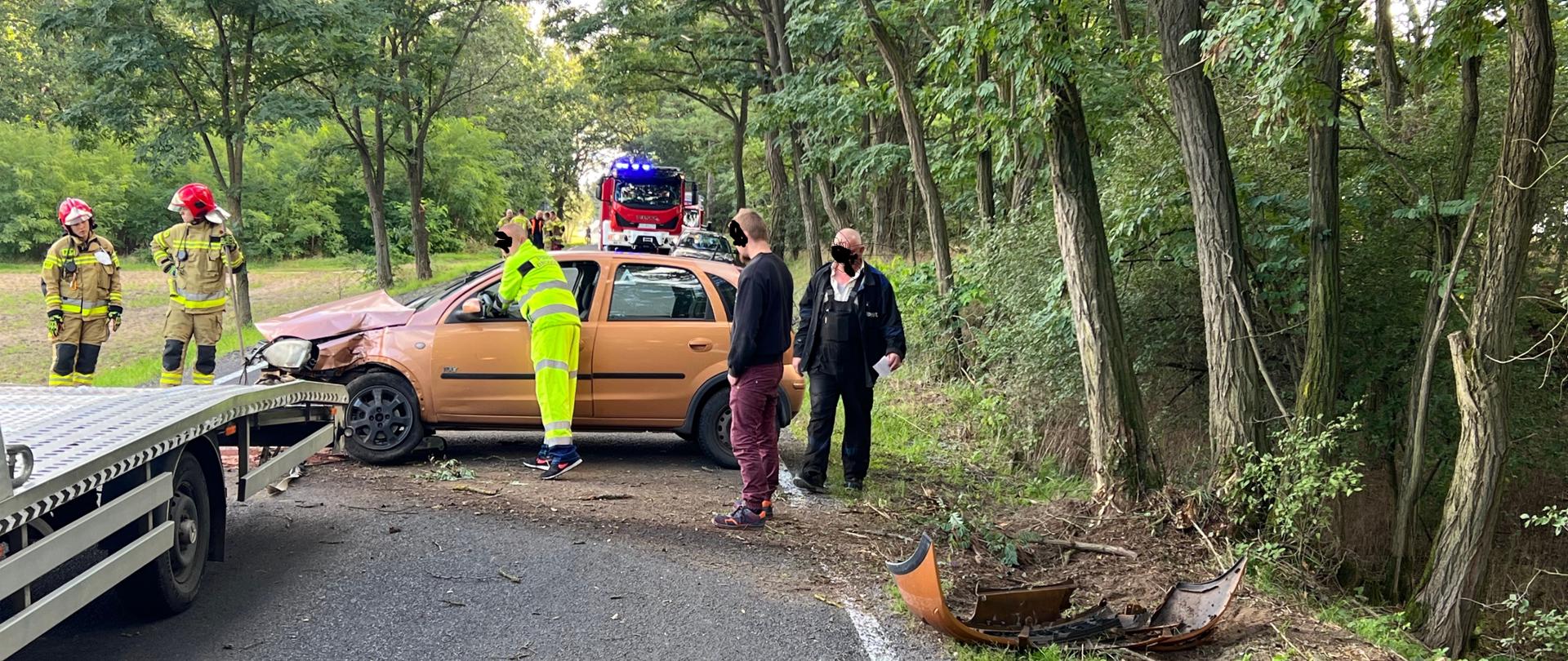 Na zdjęciu rozbity samochód osobowy przy którym jest w zielonym ubraniu pracownik pomocy drogowej obok stoją osoby postronne z przodu 2 strażaków. z lewej strony zdjęcia widać lawetę a za uszkodzonym pojazdem stoi samochód ratowniczo - gaśniczy z włączonymi sygnałami świetlnymi