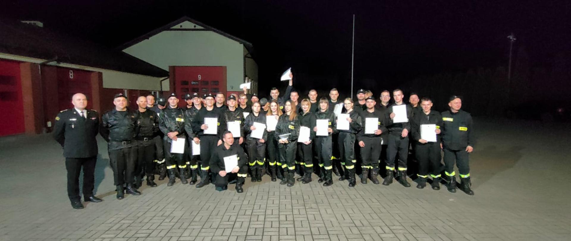 Strażacy stoją na placu na polbruku. W rękach trzymają zaświadczenia potwierdzające zdanie egzaminu. Po lewej stronie zdjęcia Komendant Powiatowy, Dowódca JRG oraz jego zastępca. Dalej strażacy po z zaświadczeniami.