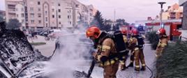Na zdjęciu widać spalony samochód w kolorze srebrnym. Nad autem obłok pary wodnej, szary. Na drugim planie budynki wielorodzinne. Zdjęcie wykonane w ciągu dnia. Na zdjęciu ratownik PSP w ubraniu specjalnym i hełmie.