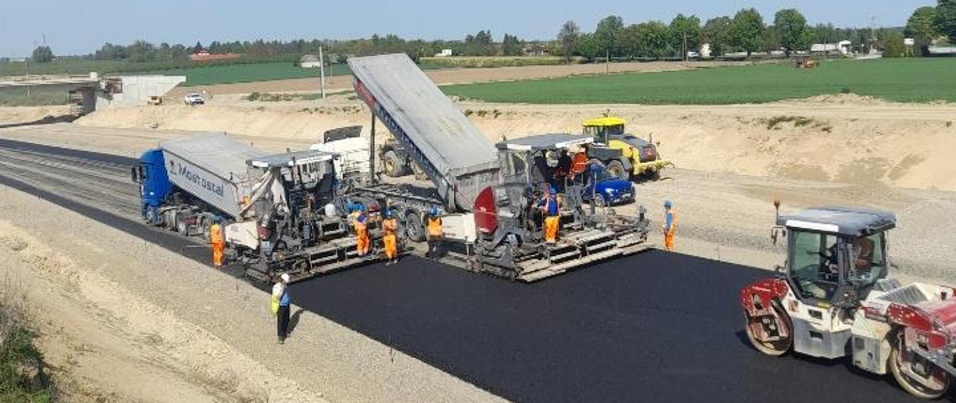 Budowa obwodnicy Opatowa - układanie podbudowy bitumicznej, na czarnej drodze maszyny dwie układarki, jeden walec, pracownicy w pomarańczowych ubraniach ochronnych 