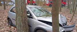 Samochód osobowy marki Opel stoi poza drogą, w lesie. Ma gnieciony pas przedni pojazdu po zderzeniu z drzewem. Jest dzień, w tle inne drzewa oraz pojazdy służb ratunkowych, na podłożu leżą liście.