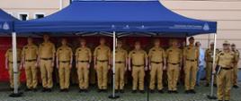 Strażacy w mundurach koloru musztardowego stoją obok siebie pod namiotem koloru niebieskiego.