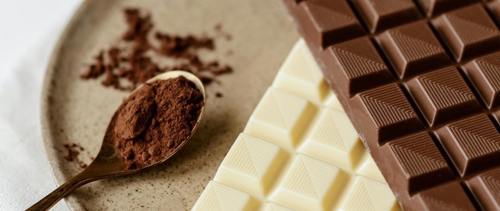 Na zdjęciu znajdują się dwie tabliczki czekolady w kolorze białym i czarnym położone na talerzyku. Obok znajduje się łyżeczka na której jest kakao.