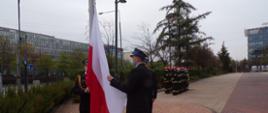 Strażacy dokonują uroczystego podniesienia Flagi Polskiej na maszt, w dalszym planie grupa strażaków ustawiona w dwuszeregu