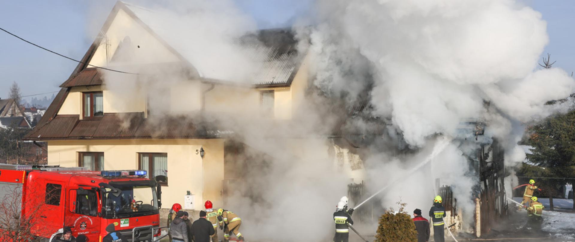 na zdjęciu palący się dom mieszkalny w miejscowości Szaflary strażacy gaszą pożar z boku budynku znajduje się czerwony samochód gaśniczy z budynku unoszą się kłęby białego i czarnego dymu działania prowadzone są w dzień