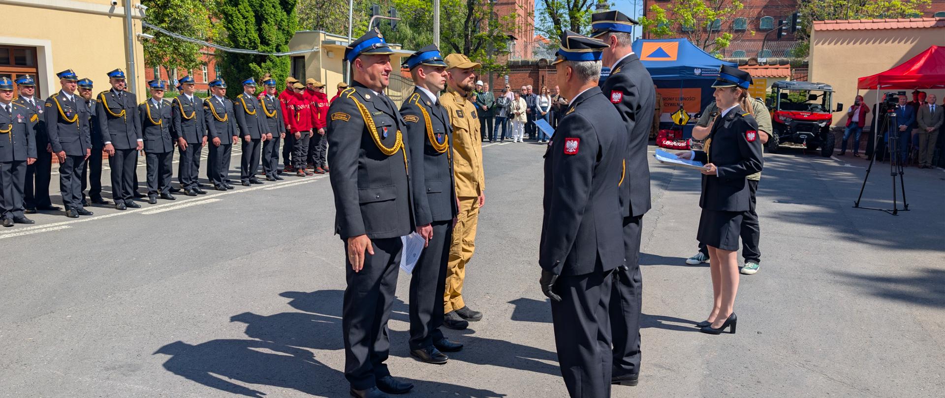 Na zdjęciu widać strażaków którzy wręczają odznaczenia. po lewej stronie osoby uzyskujący oznaczenie po prawej komendanci wręczający odznaczenia. Wszyscy w umundurowaniu galowym. W tle widać strażaków w szyku uroczystym. 