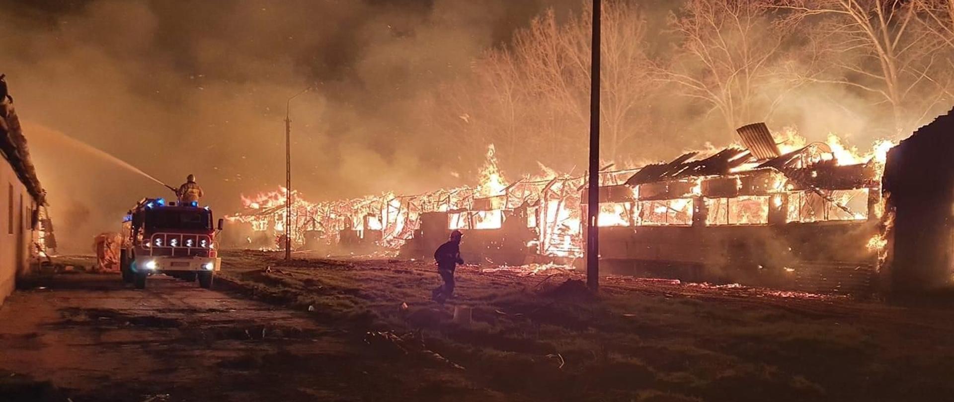 Pożar dwóch budynków gospodarczych. Pomiędzy nimi znajduje się samochód strażacki i strażacy którzy prowadzą akcję gaśniczą jednego z nich. Pożar jest w fazie bardzo rozwiniętej. Panuje nocy.
