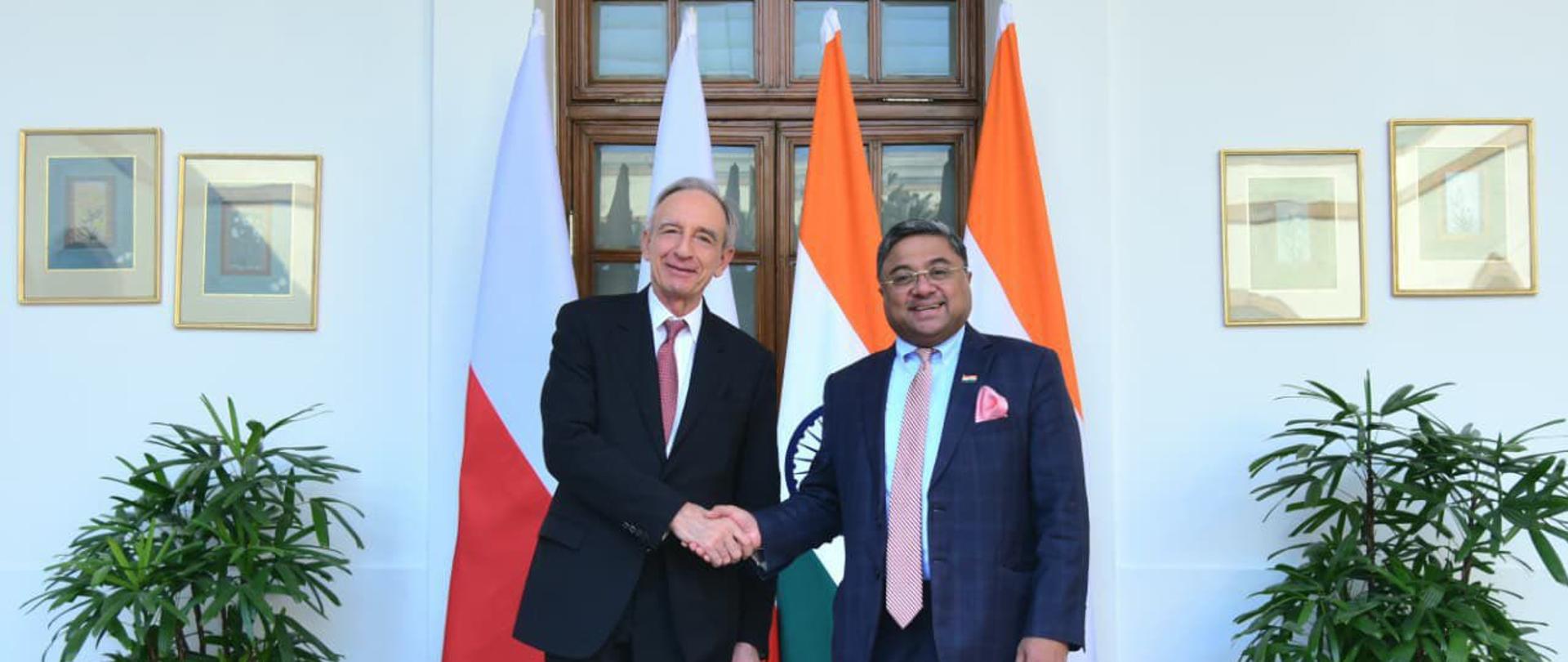 Minister Władysław Teofil Bartoszewski uściska dłoń swojemu indyjskiemu odpowiednikowi Sibi George'owi na tle flag Polski i Indii