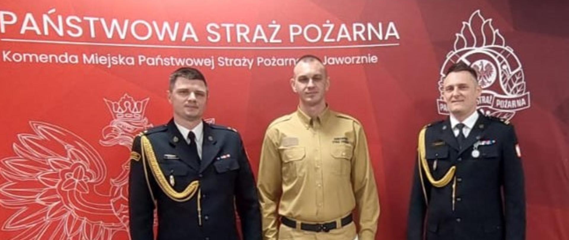 Trzech funkcjonariuszy Państwowej Straży Pożarnej pozuje do pamiątkowego zdjęcia przed ścianką Komendy Miejskiej PSP w Jaworznie; nowo przyjęty funkcjonariusz stoi pośrodku.