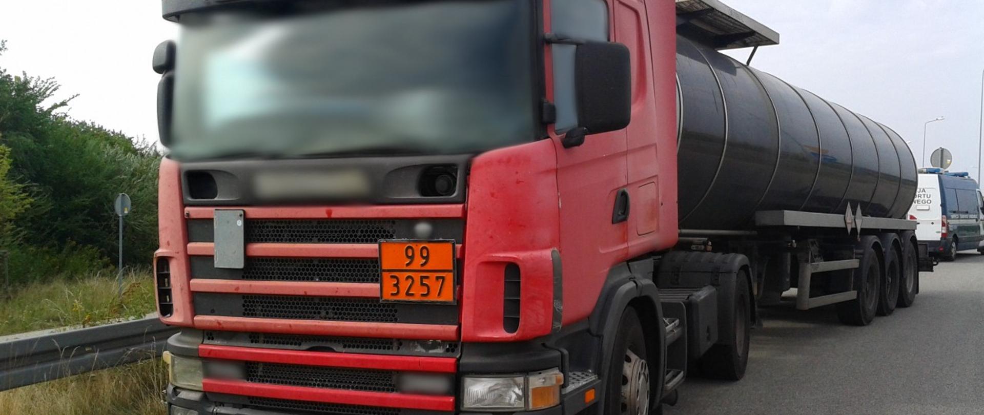 Na pierwszym planie: przód zatrzymanej do kontroli drogowej ciężarówki oznaczonej tablicą pomarańczowej barwy (ADR). Z tyłu zespołu pojazdów (ciągnika siodłowego z naczepą) stoi oznakowany furgon mazowieckiej ITD.