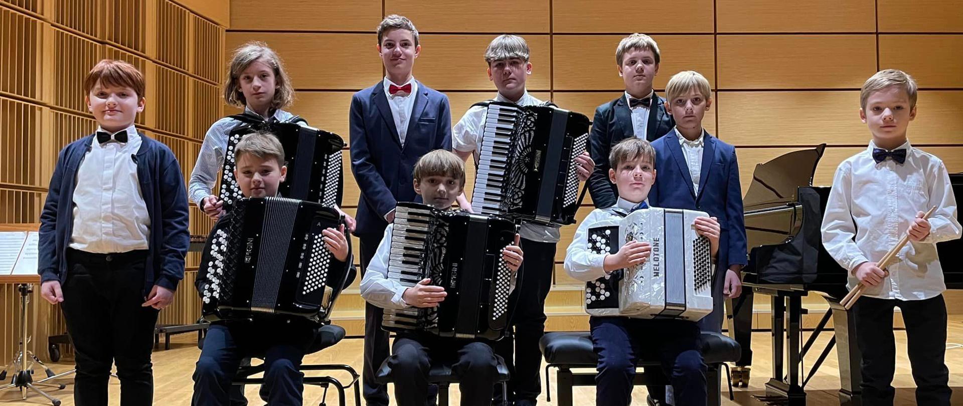 Uczniowie stojący oraz siedzący w dwóch rzędach na scenie sali koncertowej. Z przodu siedzą akordeoniści trzymający instrumenty oraz uczeń trzymający pałeczki. Z tyłu dwóch uczniów trzyma akordeony.
