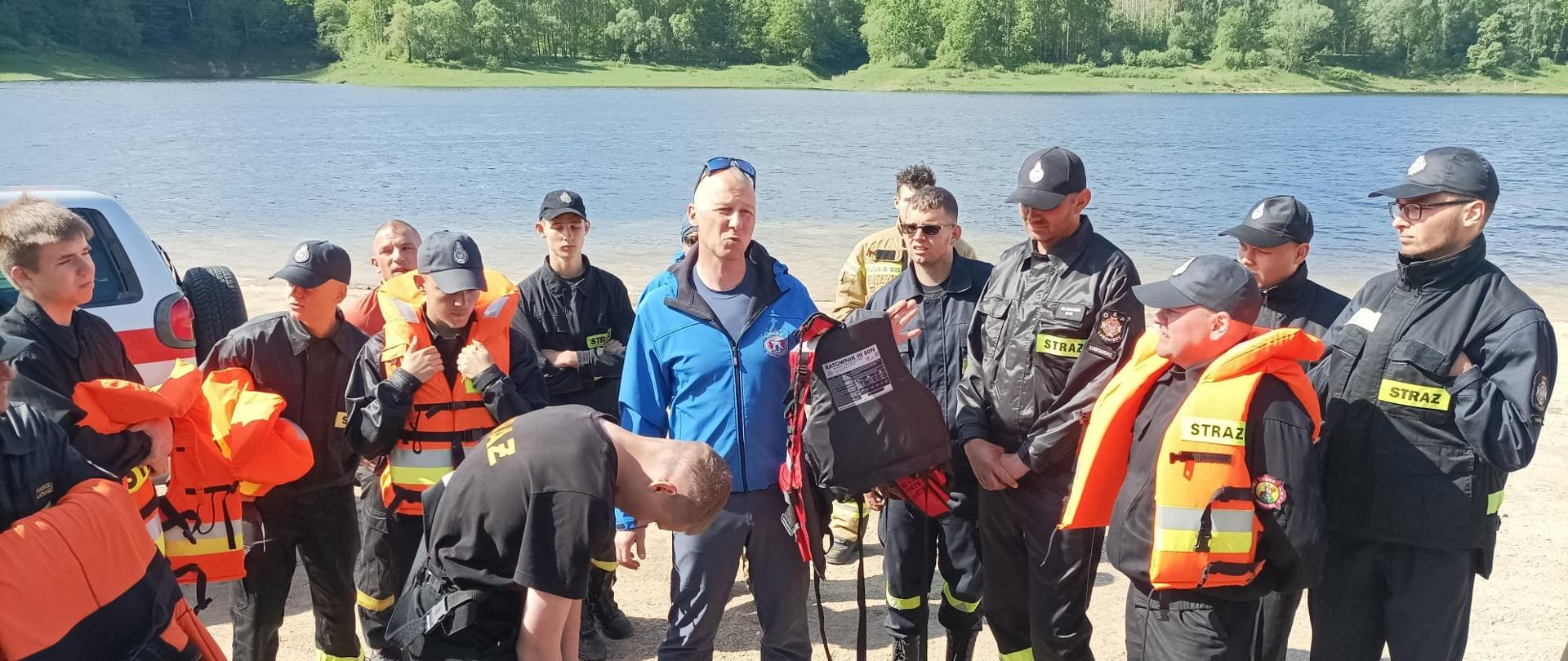 Na zdjęciu widać kilkunastu strażaków znajdujących się na plaży przed wodą. Zdjęcie przedstawia szkolenie dla strażaków ochotników. Strażak instruktor znajdujący się na środku zdjęcia w bluzie koloru niebieskiego trzyma kamizelkę ratunkową, W tle widać wodę. Zdjęcie wykonane w dzień podczas ćwiczeń,