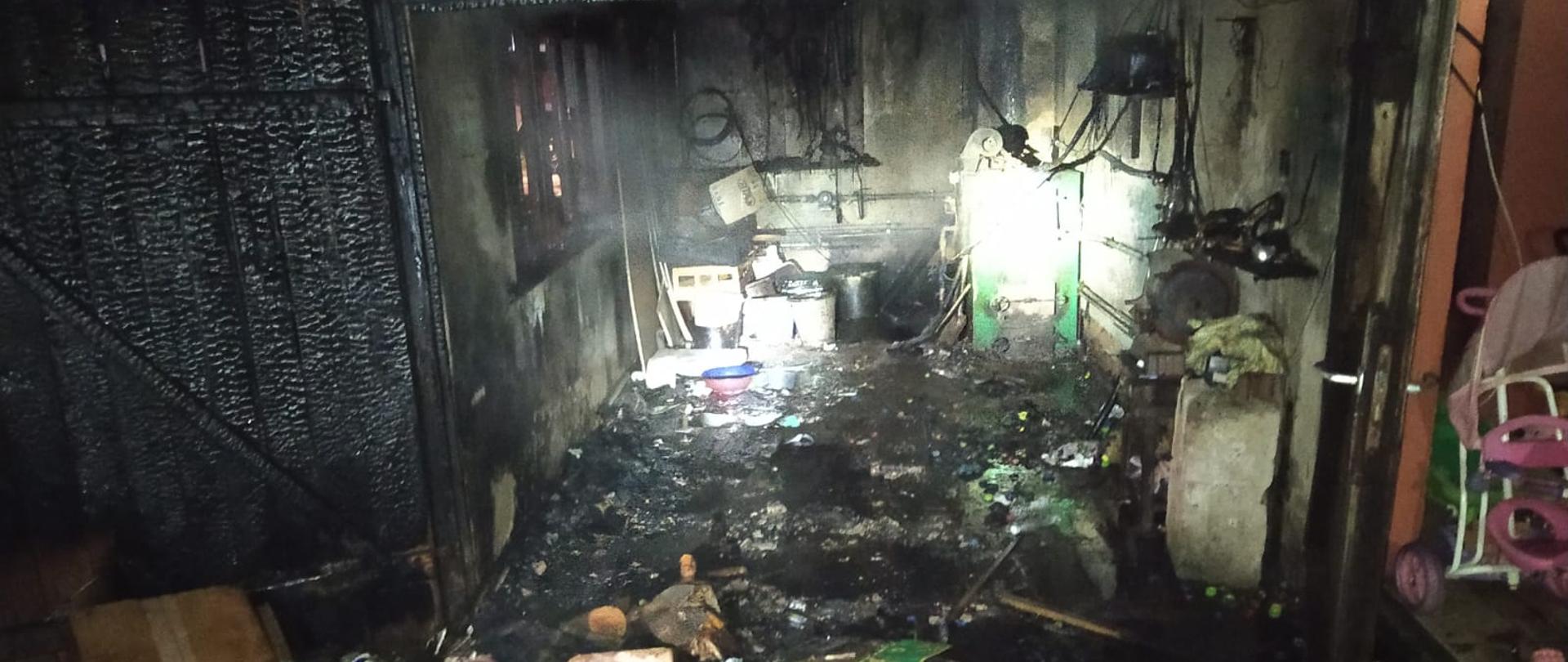 Wnętrze garażu z widocznymi elementami spalonymi i osmolonymi. Na posadzce porozrzucane elementy wyposażenia garażu. W głębi widoczny piec na paliwo stałe.