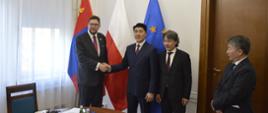 Pod ścianą sali stoi wiceminister Szeptycki, podaje rękę mężczyźnie w garniturze, obok stoi dwóch mężczyzn, za nimi flagi Polski, Mongolii i UE.