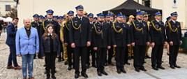 W rzędach stoją wyróżnione osoby strażacy ubrani w mundury osoby cywilne w garnitury, na klapach mają przyczepione otrzymane medale. Z tyłu widać kolejne rzędy odznaczonych i awansowanych strażaków również w mundurach galowych. W tle piaskowy budynek