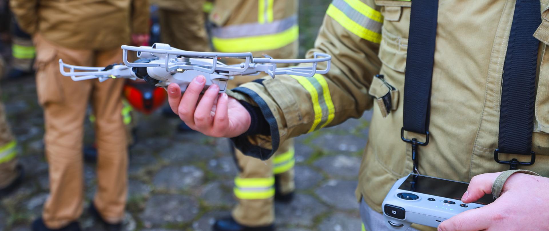 Zbliżenie na szarego drona, który jest trzymany w ręku przez strażaka. Po prawej stronie zdjęcia kontroler do tego drona. 
