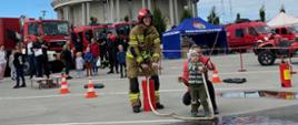 Na pierwszym planie strażak w umundurowaniu bojowym, który pompuje wodę hydronetką. Na końcu linii wężowej mały chłopiec z mamą. W tle widać kościół i ekspozycję festynową pojazdów strażackich. 