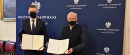 Zdjęcie przedstawia dwóch elegancko ubranych mężczyzn, trzymających w rękach umowy. Pozują oni na tle granatowej ścianki wizerunkowej Wojewody Mazowieckiego, opatrzonej kilkoma logotypami Wojewody Mazowieckiego (stanowiącymi Godło Polski oraz napis "Wojewoda Mazowiecki").