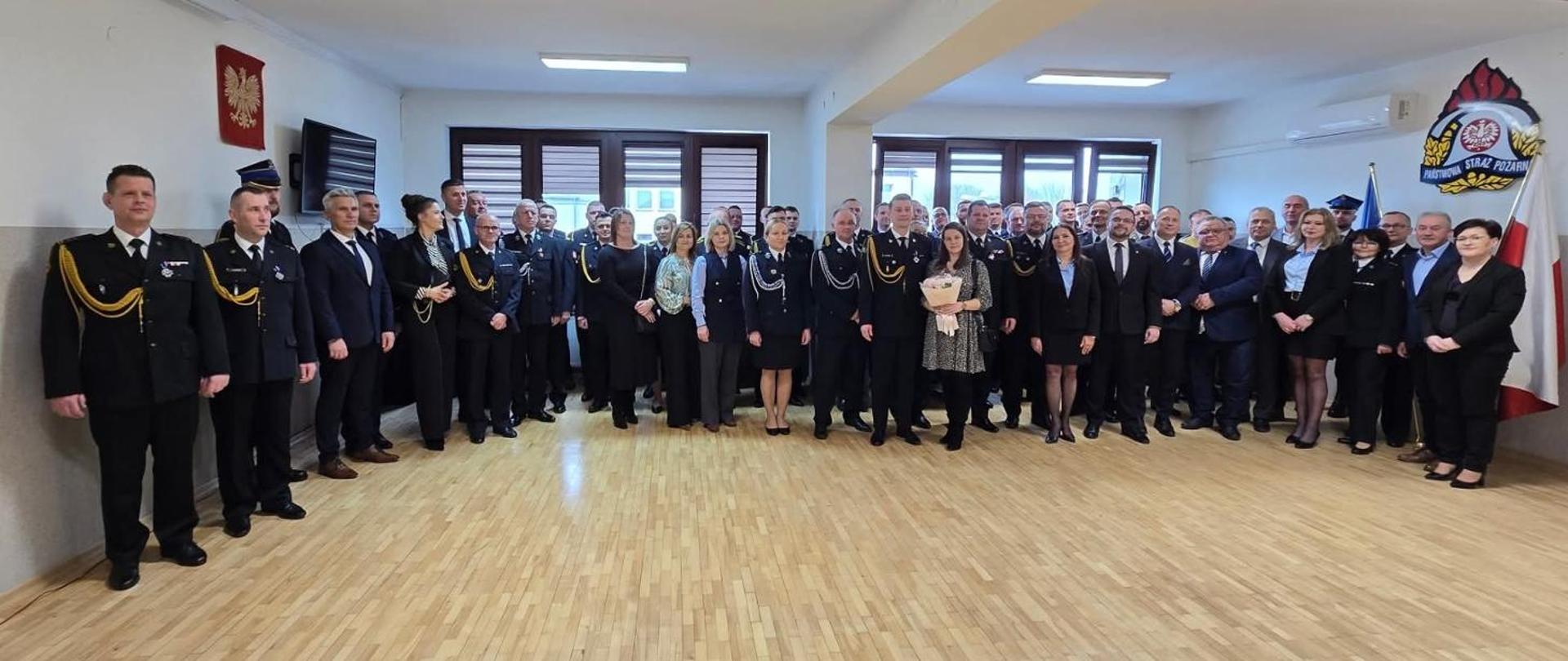Panoramiczne zdjęcie grupowe wykonane na zakończenie lub w ważnym momencie uroczystości.
Uczestnicy: W sali zgromadzonych jest kilkadziesiąt osób – strażacy w mundurach galowych i służbowych oraz osoby cywilne (kobiety i mężczyźni).
Centralny punkt: W środku grupy stoi kobieta z bukietem kwiatów oraz strażacy pełniący główne role w uroczystości.
Otoczenie: Grupa wypełnia niemal całą szerokość sali. Po lewej stronie na ścianie wisi godło Polski, a po prawej emblemat Państwowej Straży Pożarnej oraz flaga państwowa.