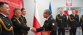 Podczas uroczystości w siedzibie Państwowej Straży Pożarnej jeden ze strażaków wręcza dokument i ściska dłoń funkcjonariuszowi stojącemu w szeregu. W tle znajdują się pozostali strażacy w galowych mundurach oraz flagi Polski i Unii Europejskiej.
