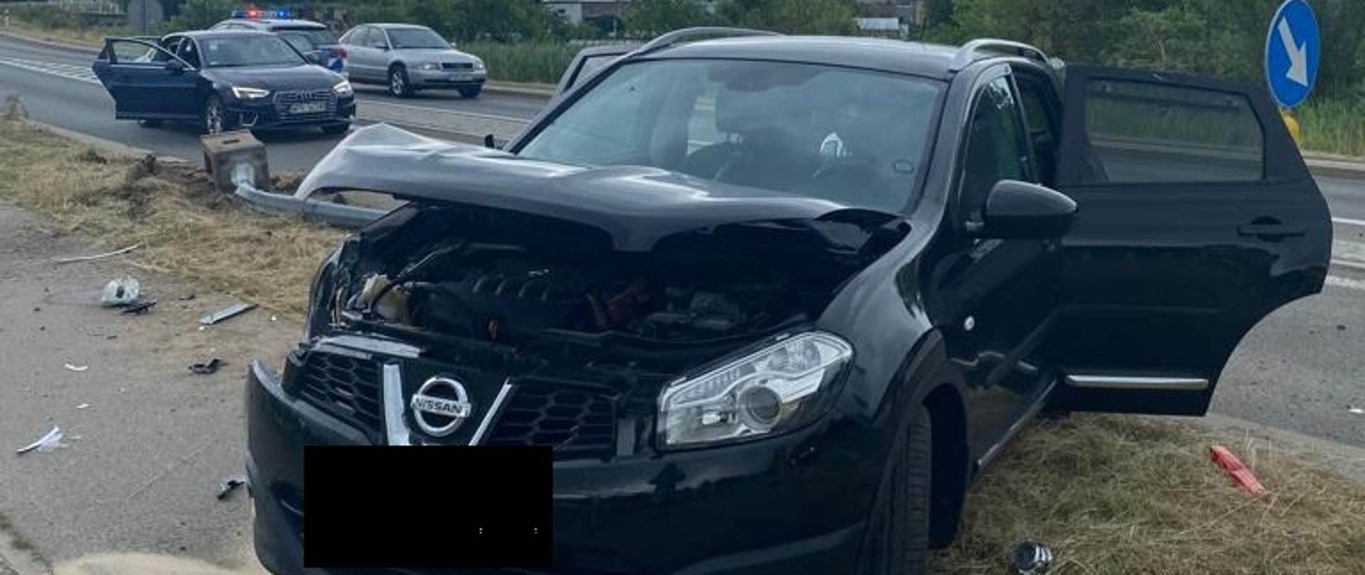 Na poboczu drogi stoi czarny samochód osobowy, który ma rozbity przód. Za nim widać wywrócona latarnie i jeszcze dwa inne samochody osobowe. 